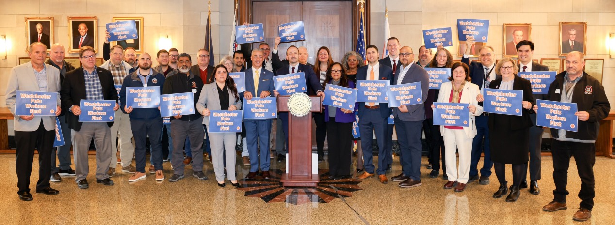 Legislators and union labor leaders stand at podium in Board rotunda holding signs.